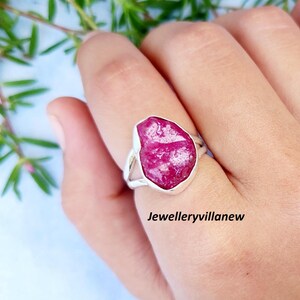 May include: A silver ring featuring a large, irregular-shaped, vibrant pink gemstone. The ring's band is a simple, polished silver, and the gemstone is the focal point. The ring is shown on a hand, with greenery in the background.