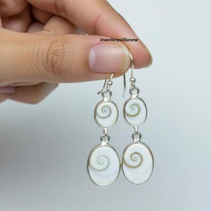 Op de afbeelding: Een paar zilveren oorhangers. Elke oorbel heeft drie ovale, witte hangers met een spiraalvormig ontwerp. De oorbellen worden door een hand tegen een lichte achtergrond gehouden. De tekst "Jewelleryvillanew" is zichtbaar.