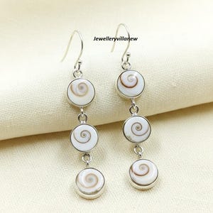 Puede incluir: Un par de pendientes colgantes con diseño de espiral. Cada pendiente presenta tres colgantes circulares blancos con un patrón de espiral marrón, conectados por eslabones plateados. Los pendientes tienen ganchos plateados. El texto "Jewelleryvillanew" es visible.