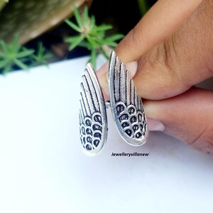 Puede incluir: Anillo plateado con dise&ntilde;o abierto, con dos elementos en forma de ala. Cada ala tiene patrones de plumas detallados y una superficie texturizada. El anillo se sostiene sobre un fondo blanco con vegetaci&oacute;n visible.