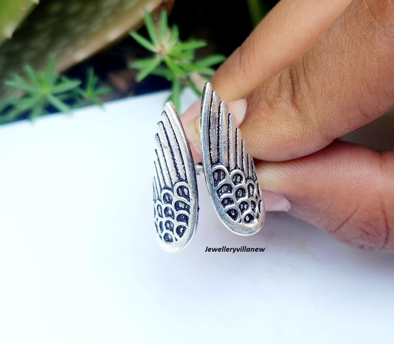 Puede incluir: Anillo plateado con dise&ntilde;o abierto, con dos elementos en forma de ala. Cada ala tiene patrones de plumas detallados y una superficie texturizada. El anillo se sostiene sobre un fondo blanco con vegetaci&oacute;n visible.