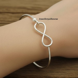 May include: A silver bangle bracelet with an infinity symbol design. The bracelet is worn on a wrist and has a simple, elegant design. The text "Jewelleryvillanew" is visible.
