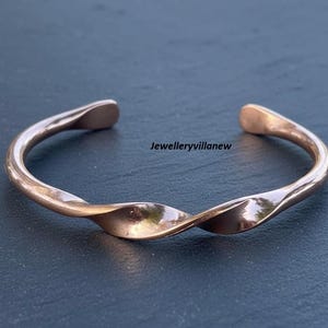 以下が含まれることがあります： 銅色の、ねじれた金属製のカフブレスレット。 オープンエンドのブレスレットは、滑らかで光沢のある仕上げと微妙な光沢があります。 デザインは中央にひねりを加え、ジュエリーに視覚的な面白さを加えています。