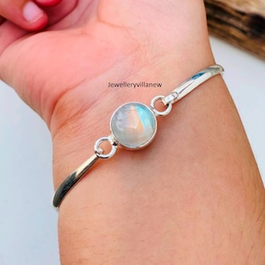 May include: A silver bangle bracelet with a round, iridescent moonstone gemstone. The bracelet is open and has a simple design.