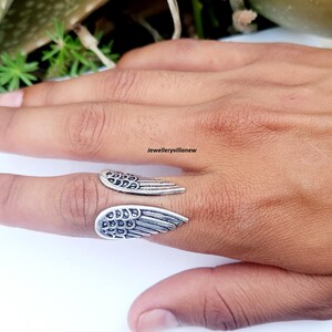Puede incluir: Un anillo plateado con dise&ntilde;o de alas. El anillo presenta dos formas de alas detalladas, una a cada lado del dedo. El anillo se lleva en un dedo, mostrando los intrincados detalles de las plumas.