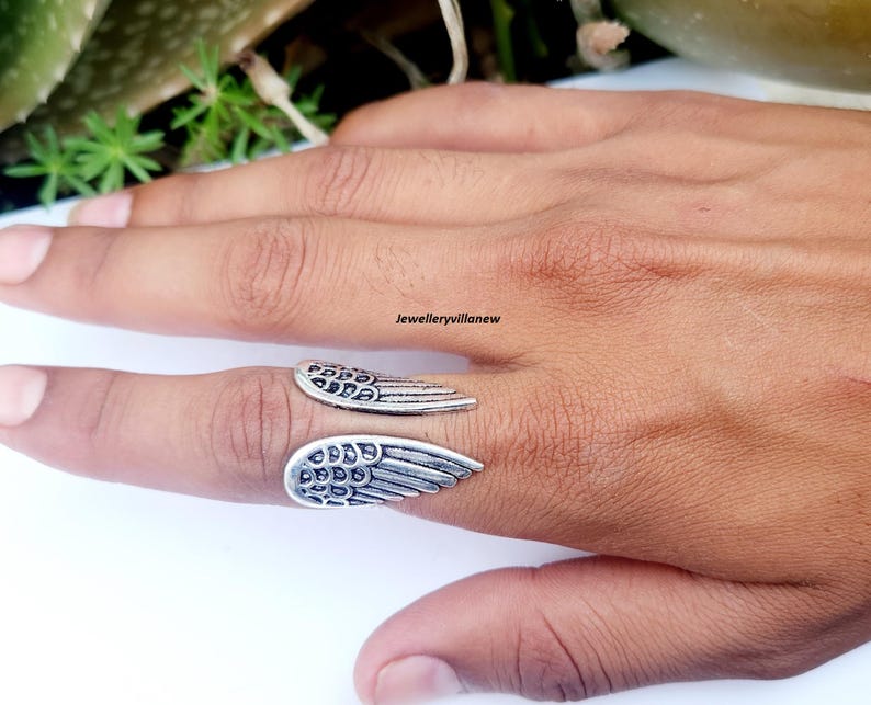 Puede incluir: Un anillo plateado con dise&ntilde;o de alas. El anillo presenta dos formas de alas detalladas, una a cada lado del dedo. El anillo se lleva en un dedo, mostrando los intrincados detalles de las plumas.