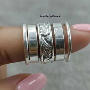 Puede incluir: Un conjunto de anillos de plata mostrados en un dedo. Los anillos tienen diseños variados, incluyendo bandas lisas y uno con un patrón floral. Los anillos están hechos de un material metálico brillante. El texto "Jewelleryvillanew" es visible.