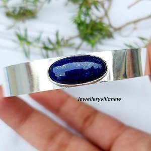 Pode incluir: Uma pulseira de punho de prata com uma grande pedra oval de lápis-lazúli azul escuro. A pedra é colocada em uma faixa de prata polida. A pulseira é exibida em um fundo macio e desfocado com vegetação.