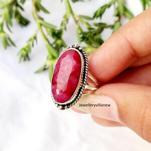 May include: A silver ring featuring a large, oval-cut ruby gemstone. The ruby is a vibrant red color and is set in a detailed silver band with a twisted rope design around the gemstone. The ring is held by a hand.