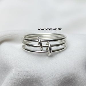 May include: A silver ring with a unique design. The ring features four bands held together by two vertical, rounded bars. The jewelry is displayed on a white, textured surface. The text "Jewelleryvillanew" is visible.
