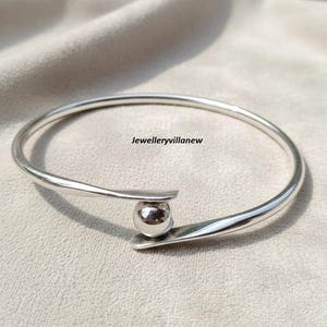 May include: A silver-colored bangle bracelet with a spherical accent. The bracelet has a minimalist design with a curved shape and a small ball in the center. The text "Jewelleryvillanew" is visible.