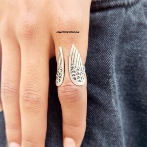 Puede incluir: Anillo plateado con un dise&ntilde;o abierto que presenta detalles en forma de alas. Las alas del anillo est&aacute;n intrincadamente dise&ntilde;adas con patrones similares a plumas. El anillo se lleva en un dedo, con el texto "Jewelleryvillanew" visible.
