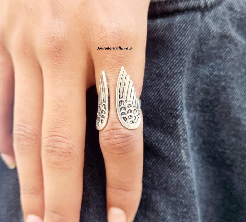 Puede incluir: Anillo plateado con un dise&ntilde;o abierto que presenta detalles en forma de alas. Las alas del anillo est&aacute;n intrincadamente dise&ntilde;adas con patrones similares a plumas. El anillo se lleva en un dedo, con el texto "Jewelleryvillanew" visible.