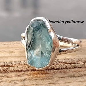 Op de afbeelding: Een zilveren ring met een grote, onregelmatig gevormde, lichtblauwe edelsteen. De ring heeft een split-schacht ontwerp. De tekst "Jewelleryvillanew" is zichtbaar.