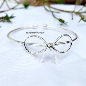 Puede incluir: Una pulsera de puño abierta de color plateado con un diseño de lazo en el centro. El lazo está detallado con un nudo y dos extremos colgantes. La pulsera tiene un acabado liso y pulido, sobre un fondo blanco. El texto "Jewelleryvillanew" es visible.