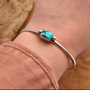 Puede incluir: Un brazalete de plata con una piedra turquesa engastada en un marco de plata. El brazalete está en la muñeca de una persona.