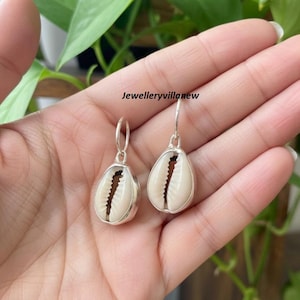 Puede incluir: Un par de pendientes de aro plateados con colgantes de concha de cauri natural. Las conchas son de color crema con una ranura marrón oscuro. El texto "Jewelleryvillanew" es visible.