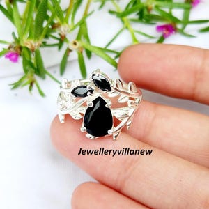 May include: A silver ring with a leaf design, featuring three black gemstones. The largest gemstone is pear-shaped, and the other two are oval. The ring is held against a blurred background of green leaves and pink flowers.