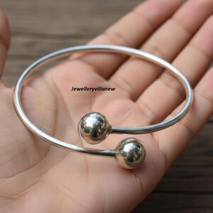 May include: A silver-colored bangle bracelet with two spherical end pieces. The bracelet is held in a hand, and the surface of the bracelet reflects light. The text "Jewelleryvillanew" is visible.