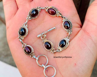 May include: A silver bracelet with a toggle clasp and six oval-shaped red gemstones. The bracelet is on a hand.