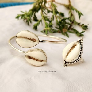 May include: A silver bangle bracelet and ring set featuring cowrie shell accents. The bracelet is open and has two cowrie shells on the front. The ring has a single cowrie shell and silver bead accents.