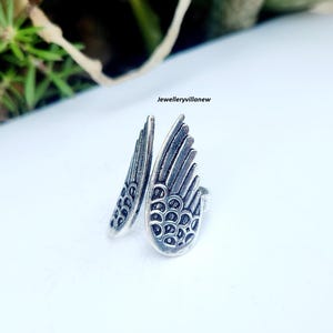 Puede incluir: Un anillo plateado con forma de dos alas estilizadas. Las alas est&aacute;n detalladas con l&iacute;neas en forma de plumas y peque&ntilde;os patrones circulares. El anillo se encuentra sobre un fondo blanco, con el texto "jewelleryvillanew" visible.