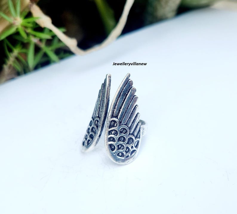 Puede incluir: Un anillo plateado con forma de dos alas estilizadas. Las alas est&aacute;n detalladas con l&iacute;neas en forma de plumas y peque&ntilde;os patrones circulares. El anillo se encuentra sobre un fondo blanco, con el texto "jewelleryvillanew" visible.