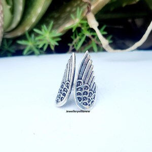 Puede incluir: Un anillo plateado con forma de alas de &aacute;ngel. El anillo presenta patrones de plumas detallados y est&aacute; colocado sobre una superficie blanca con follaje verde en el fondo. El texto "Jewelleryvillanew" es visible.