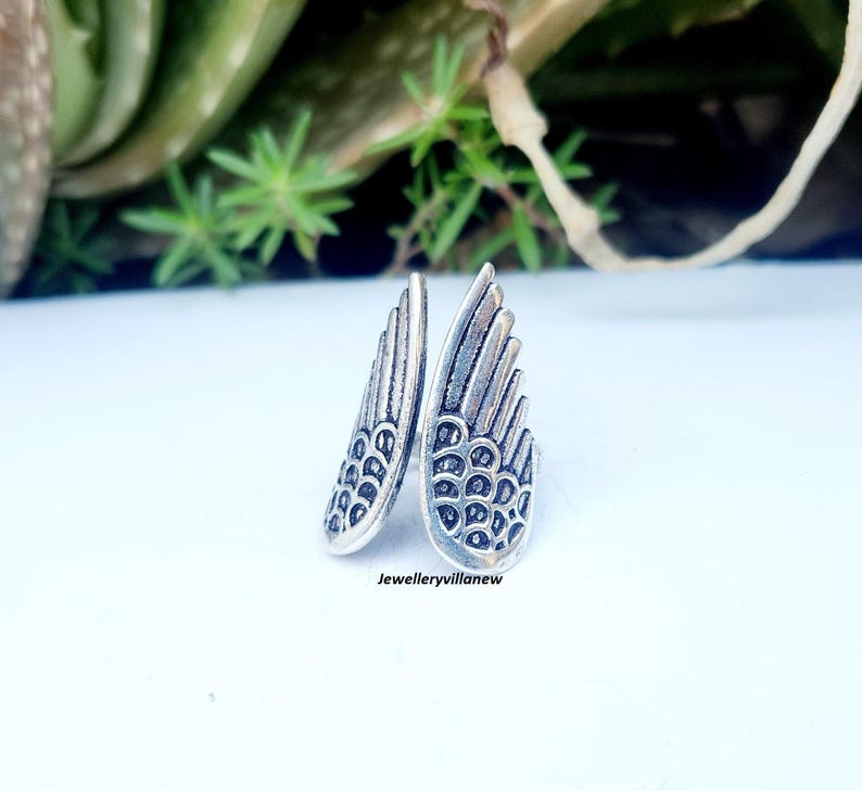 Puede incluir: Un anillo plateado con forma de alas de &aacute;ngel. El anillo presenta patrones de plumas detallados y est&aacute; colocado sobre una superficie blanca con follaje verde en el fondo. El texto "Jewelleryvillanew" es visible.
