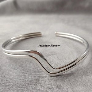 Può includere: Un bracciale a polsino color argento con un design a forma di V. Il bracciale ha una finitura liscia e lucida ed è aperto alle estremità. Il testo "Jewelleryvillanew" è visibile sulla superficie.