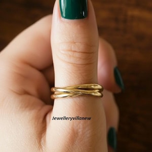 May include: A gold-colored ring with a crisscross design is worn on a finger with dark green nail polish. The ring is smooth and shiny, and the text "Jewelleryvillanew" is visible.