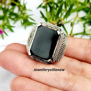 May include: A silver-toned ring with a large, rectangular black stone. The ring has a textured, detailed band and is displayed on a hand. The text "Jewelleryvillanew" is visible.