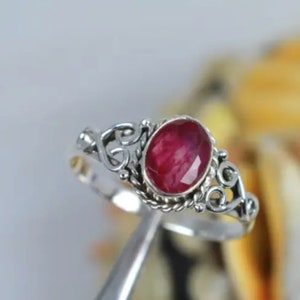 Może przedstawiać: Srebrny pierścionek z czerwonym rubinem osadzonym w delikatnym, ozdobnym wzorze.