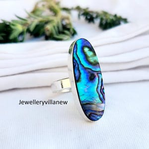Puede incluir: Un anillo de plata con una gran piedra ovalada de concha de abulón. La concha tiene colores iridiscentes azules, verdes y morados.
