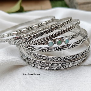 Puede incluir: Varios brazaletes plateados con intrincados diseños florales y de hojas. Algunos brazaletes presentan pequeñas piedras redondas y opalescentes. Los brazaletes están apilados sobre una tela blanca. El texto "Jewelleryvillanew" es visible.