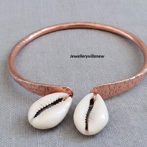 Könnte beinhalten: Kupferfarbene Halskette mit gehämmertem Muster. Zwei weiße Kaurischnecken hängen von der Vorderseite der Halskette. Der Text "Jewelleryvillanew" ist im Bild zu sehen.