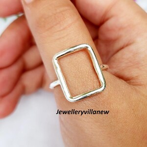 May include: A silver ring with a rectangular open design. The ring is worn on a finger, showcasing the geometric shape. The jewelry is simple and modern, with a polished finish. The text "Jewelleryvillanew" is visible.