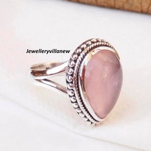 May include: A silver ring with a large, oval rose quartz gemstone. The ring has a split shank design and a beaded border around the gemstone. The text "Jewelleryvillanew" is visible on the left side of the ring.