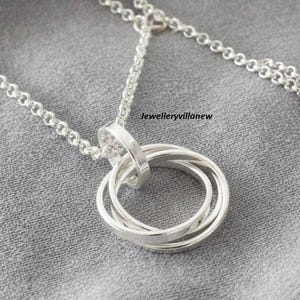 Peut inclure: Un collier argenté avec un pendentif composé de quatre anneaux entrelacés. La chaîne est fine. Le bijou est présenté sur un fond en tissu gris. Le texte "jewelleryvillanew" est visible.