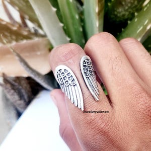 Puede incluir: Un anillo plateado con un dise&ntilde;o abierto, con dos formas de alas estilizadas. El anillo se lleva en un dedo, con las alas extendi&eacute;ndose hacia afuera. El anillo est&aacute; detallado con patrones de plumas.