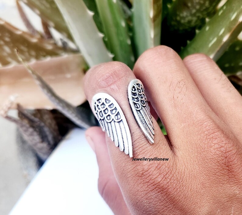 Puede incluir: Un anillo plateado con un dise&ntilde;o abierto, con dos formas de alas estilizadas. El anillo se lleva en un dedo, con las alas extendi&eacute;ndose hacia afuera. El anillo est&aacute; detallado con patrones de plumas.