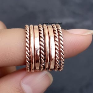 Puede incluir: Una colección de anillos de color cobre, algunos con un diseño de cuerda trenzada, apilados en un dedo. Los anillos tienen un acabado pulido y varían en ancho. El fondo es un gris oscuro borroso.