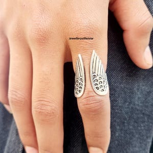 Puede incluir: Un anillo plateado con dise&ntilde;o de alas. El anillo tiene un dise&ntilde;o abierto, con cada ala detallada con l&iacute;neas y patrones grabados. El anillo se lleva en un dedo, destacando el intrincado dise&ntilde;o. Las alas del anillo est&aacute;n detalladas.