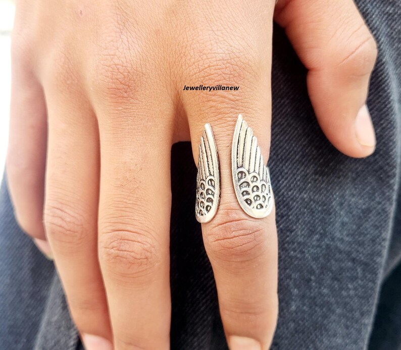 Puede incluir: Un anillo plateado con dise&ntilde;o de alas. El anillo tiene un dise&ntilde;o abierto, con cada ala detallada con l&iacute;neas y patrones grabados. El anillo se lleva en un dedo, destacando el intrincado dise&ntilde;o. Las alas del anillo est&aacute;n detalladas.