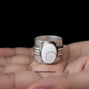 May include: A silver ring with an oval, white stone featuring a spiral design. The ring has a textured band with floral patterns and the text "Jewelleryvillanew".