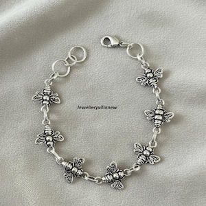 Könnte beinhalten: Ein silberfarbenes Armband mit detaillierten Bienen-Anhängern. Das Armband hat einen Karabinerverschluss und wird auf einer neutralen Oberfläche präsentiert. Der Text "Jewelleryvillanew" ist sichtbar.