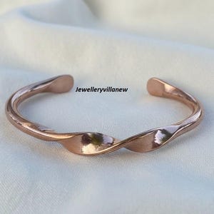 以下が含まれることがあります： ねじれたデザインのローズゴールドカラーのカフブレスレット。オープンエンドのブレスレットは光沢のある金属製で、「Jewelleryvillanew」の文字が入っています。ブレスレットは白い表面に表示されています。