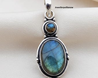 Könnte beinhalten: Ein silberner Anhänger mit einem ovalen, irisierenden blauen Edelstein. Der Anhänger hat einen kleineren, runden Edelstein über dem ovalen Stein. Der Schmuck ist vor einem weißen Hintergrund platziert. Der Text auf der rechten Seite des Bildes lautet "Jewelleryvillanew".