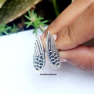 Puede incluir: Un anillo plateado con dise&ntilde;o de alas. El anillo presenta dos alas abiertas, cada una con patrones de plumas detallados y una superficie texturizada. El anillo se sostiene contra un fondo borroso de follaje verde.