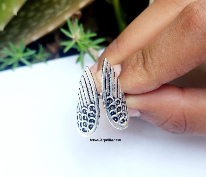 Puede incluir: Un anillo plateado con dise&ntilde;o de alas. El anillo presenta dos alas abiertas, cada una con patrones de plumas detallados y una superficie texturizada. El anillo se sostiene contra un fondo borroso de follaje verde.